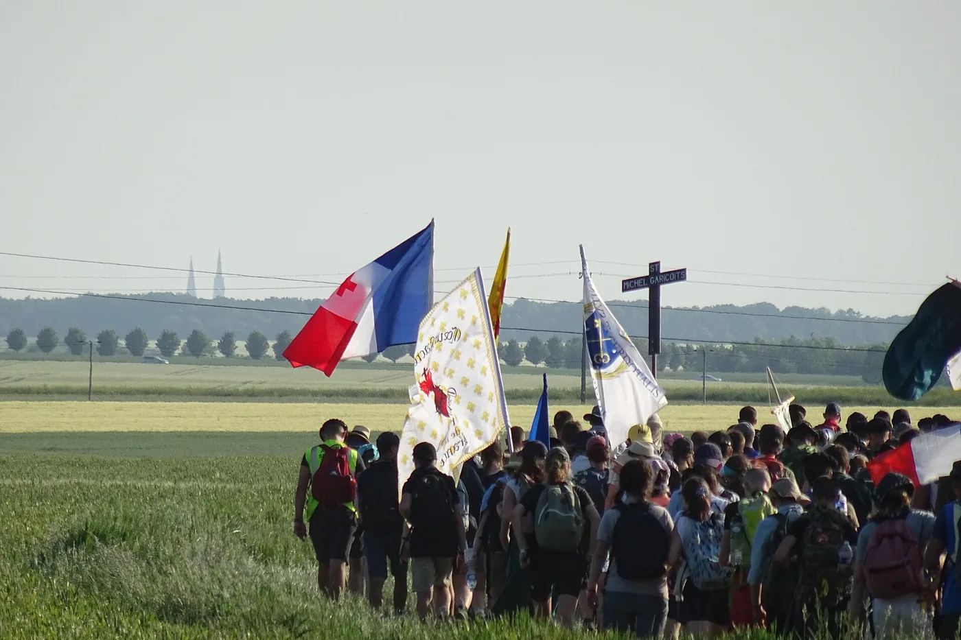 Coluna de peregrinos de costas, com estandartes, bandeiras e a cruz de um capítulo, atravessando um trigal verde. No horizonte, as duas flechas da catedral de Chartres.
